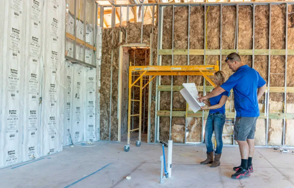 Man and woman standing in a new home holding blueprints in hand during rough in phase with framing exposed