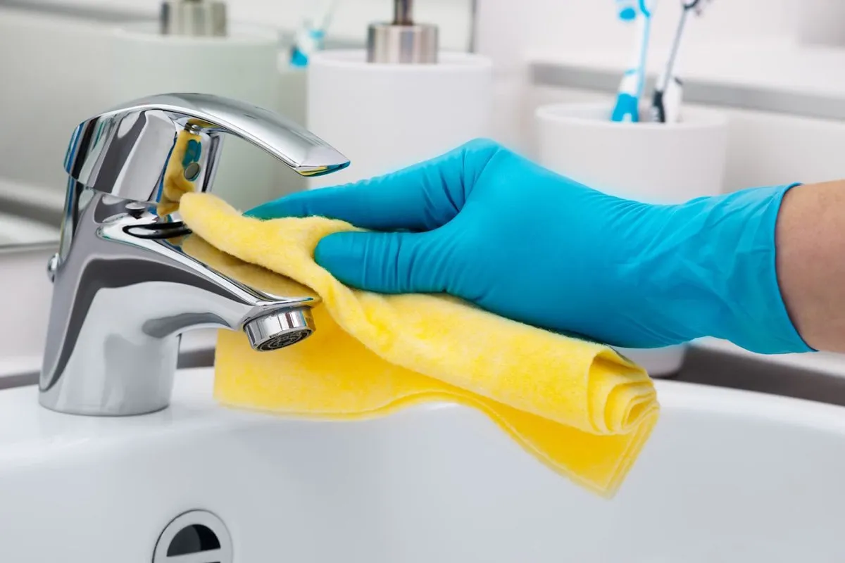 Close up picture of a hand with blue rubber glove cleaning a chrome faucet with a yellow towel