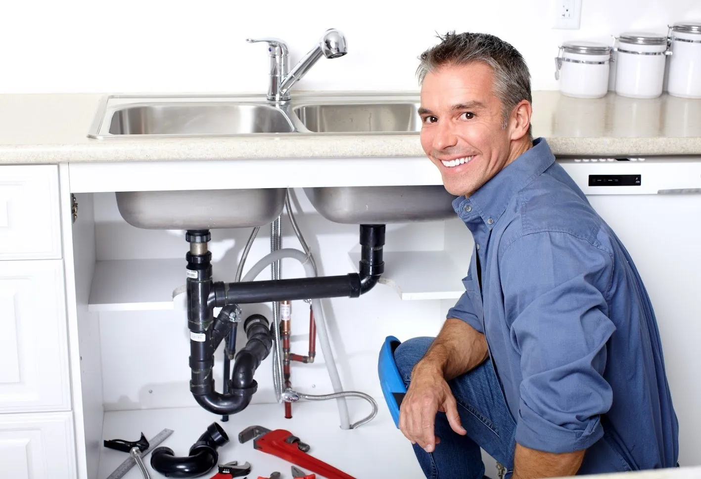 Happy plumber working on kitchen sink with cabinet open showing sink drain